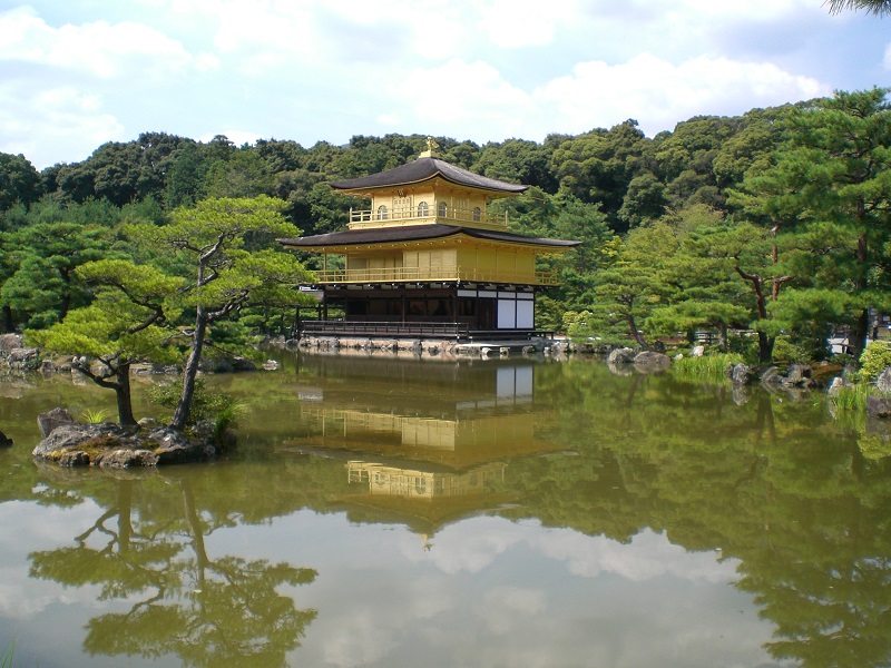 middle: three storied yellow and black building, a line of trees across the rest of the midground; foreground: dark green water with reflections and a small island; background: cloudy sky