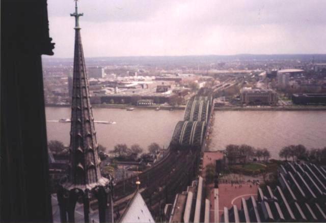edge and spire of Köln Cathedral on left, view of curvy-topped bridge over Rhein River
