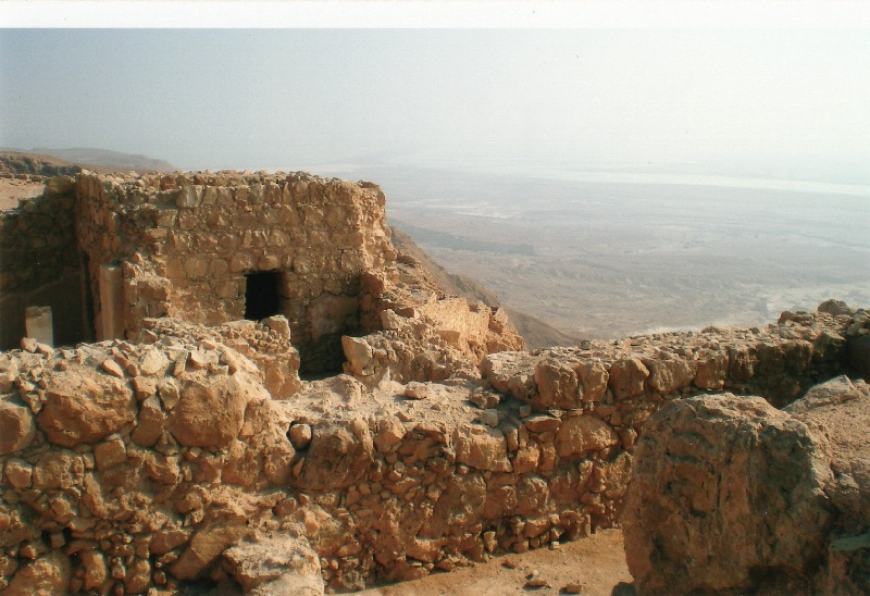 foreground: crumbled fortress walls; no mid-ground; background below the sky: hazy plains far below, with the Dead Sea in the distance