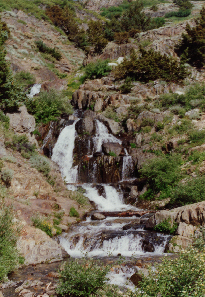 cascading white water in a zig-zag with green bushes and gray rocks to its left and right