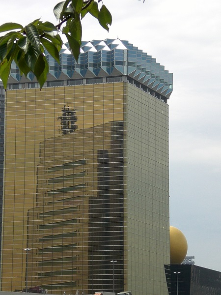 foreground top: leafy green branch; midground: modern skyscraper with bevelled articulations near the roof and the reflection of another building in its surface; background: hazy sky