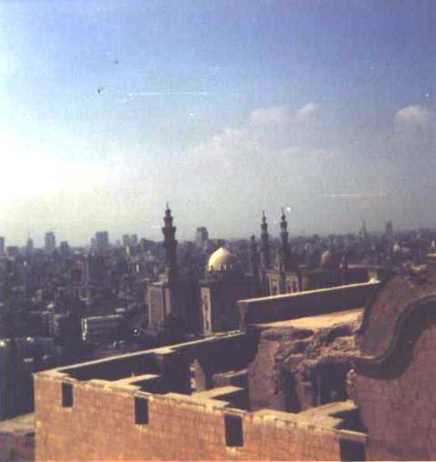 ruined wall in foreground, mosque with dome and minarets in mid-ground; modern buildings extending to horizon in background
