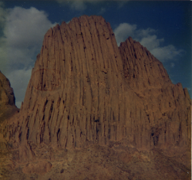 Yellow vertical rock mountain, deeply creased; foreground rock rubble of the same color; background: deep blue sky with some clouds