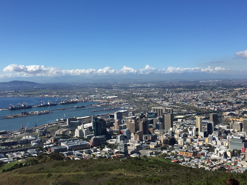 foreground: green grass on hilltop; midground: skyscrapers (mostly gray); background from mid to deep back: low rise buildings on right, piers on water on left; hazy urban to the right, blue gray mountain on left; row of clouds; blue sky