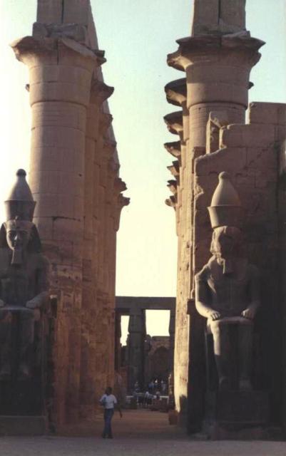 foreground: two seated statues with bucket-cone crowns; mid-ground: double wall seen edge on with columns, top out of the picture; background: much lower colonnade with entablature