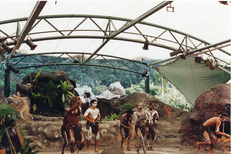 painted dancers in an open air theater bounded by painted rocks and metal hoops, with green forested hills visible in the background