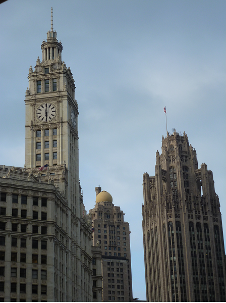 left foreground: gray building with a clock tower; right foreground: gray building with Gothic spires; rest: hazy blue sky